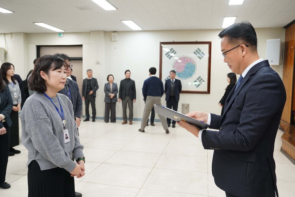 2026년 상반기 4급이하 정기인사로 정장을 입은 남성이 실내 행사장에서 한 여성에게 상장을 전달하며 내용을 읽고 있고, 여성은 회색 니트 스웨터를 입고 사원증을 착용한 채 두 손을 모은 상태로 서 있으며 뒤쪽으로 참석자들이 줄지어 서서 행사를 지켜보고 있다.