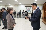 2026년 상반기 4급이하 정기인사로 정장을 입은 남성이 실내 행사장에서 한 여성에게 상장을 전달하며 내용을 읽고 있고, 여성은 갈색 파마머리에 갈색 자켓을 입고 사원증을 착용한 채 두 손을 모은 상태로 서 있으며 뒤쪽으로 참석자들이 줄지어 서서 행사를 지켜보고 있다.